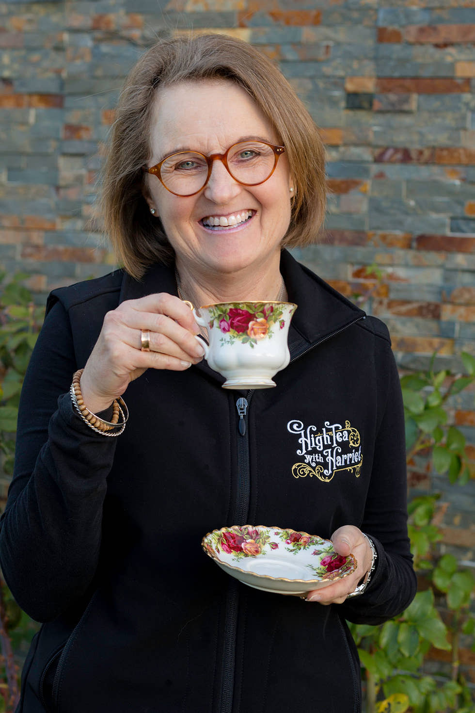 High Tea with Harriet’s former owner Denise holding a floral teacup and saucer in front of a rustic brick wall.