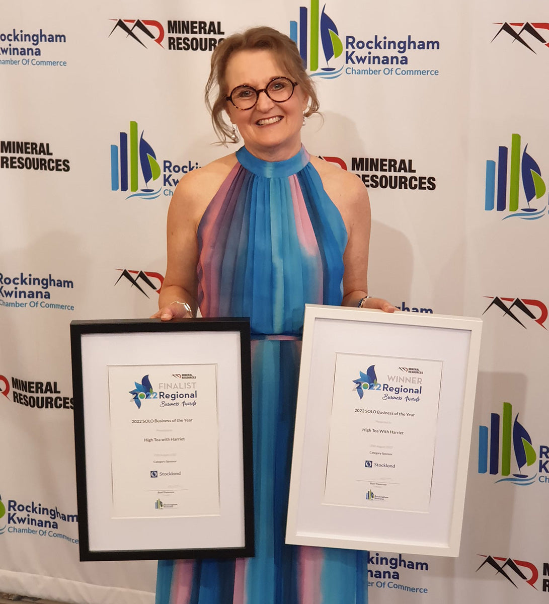 Denise smiling while holding two framed awards at the Rockingham Kwinana Chamber of Commerce event backdrop.
